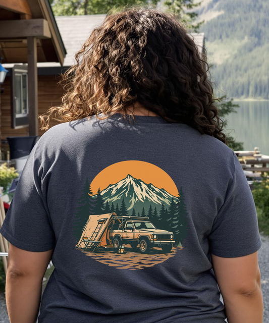 Person wearing a t-shirt with a mountain and tent graphic, standing outdoors near a cabin and lake.