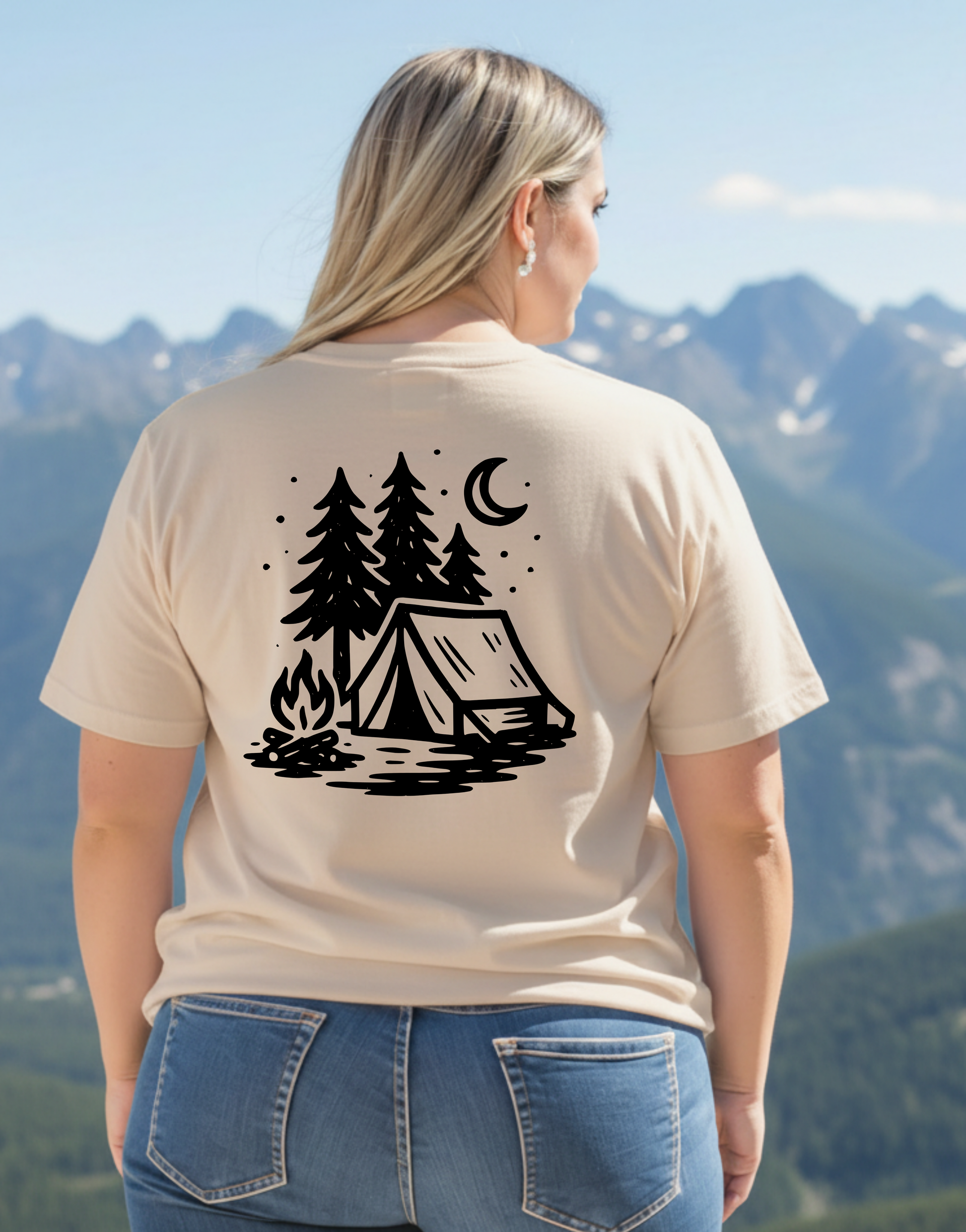 Person wearing a beige t-shirt with a camping design, standing in front of mountains.