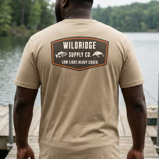 Man wearing a beige t-shirt with Wildridge Supply Co. logo by a lake.